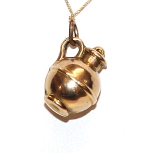 May include: A gold-toned metal charm shaped like a small, rounded jug or pitcher with a handle and a short neck. The charm is suspended from a thin, delicate gold-toned chain. The surface of the metal is smooth and reflective with a warm, metallic finish.