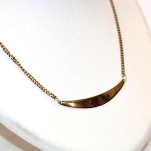 May include: A gold-coloured necklace with a curved bar pendant. The necklace has a delicate chain and a polished, crescent-shaped pendant. The jewellery is displayed on a white surface, highlighting the warm metallic tones.