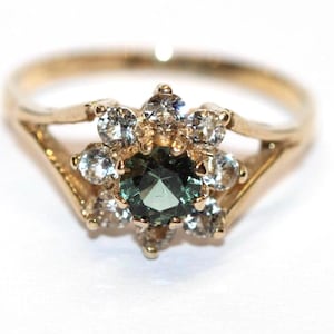 Op de afbeelding: Een goudkleurige ring met een centrale, donkergroene edelsteen, omringd door kleinere, heldere edelstenen in een bloemmotief. De band van de ring heeft een delicaat, gesplitst ontwerp. De ring staat tegen een witte achtergrond.
