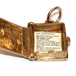 May include: A gold-coloured charm in the shape of an open book. The charm has a small ring at the top for attachment. Inside the book, a printed page displays the text of "The Lord's Prayer". The cover of the book reads "THIS BOOK BELONGS".
