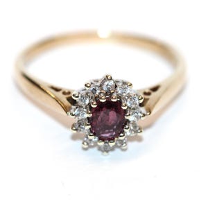 Könnte beinhalten: Goldener Ring mit einem ovalen Rubin in der Mitte, umgeben von einem Kranz aus kleinen Diamanten. Der Ring hat ein klassisches Design und ist auf einem weißen Hintergrund abgebildet, der ihn hervorhebt.