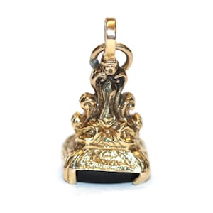 May include: A gold-toned metal wax seal stamp pendant featuring an ornate, sculpted handle with scrolling details and a rectangular base. The bottom of the seal is set with a flat, opaque black stone insert, and a small jump ring is attached to the top.