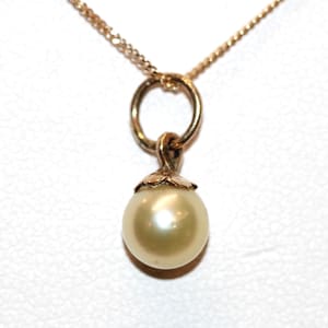 Könnte beinhalten: Eine goldfarbene Halskette mit einem einzelnen runden, cremefarbenen Perlenanhänger. Die Perle ist mit einer kleinen goldfarbenen Metallkappe mit blattartigem Design befestigt, die an einem runden goldfarbenen Bindering und einer feinen Kette hängt.