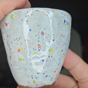 May include: A speckled ceramic cup with a light gray exterior and a brown base. The cup is adorned with colorful specks of red, yellow, blue, and green. The cup is held in a hand.