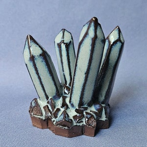 May include: A ceramic sculpture of a cluster of crystals. The crystals are a light blue color with dark brown stripes. The base of the sculpture is made of brown ceramic and has a rough texture.