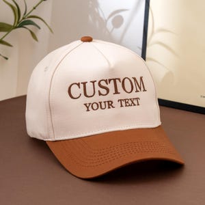 Puede incluir: Una gorra de béisbol de color crema con visera marrón y un botón marrón en la parte superior. El panel frontal presenta el texto bordado en marrón CUSTOM YOUR TEXT. La gorra tiene una corona estructurada y costuras en la visera.