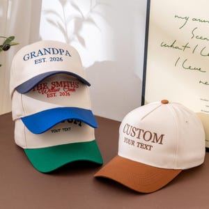 Puede incluir: Una pila de tres gorras de béisbol color hueso con viseras azul marino, azul y verde, con texto bordado que incluye GRANDPA EST. 2026 y THE SMITHS Willow Creek EST. 2026. Al lado, una gorra color hueso con visera marrón muestra CUSTOM YOUR TEXT.