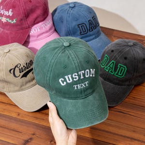May include: A collection of baseball caps in various colors, including green, blue, pink, and tan. Each cap features embroidered text, such as "CUSTOM TEXT," "DAD," and "Sarah." The caps have a vintage, worn look.