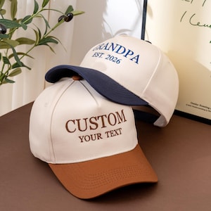 Puede incluir: Dos gorras de béisbol con coronas color blanco hueso. La gorra superior tiene una visera azul marino y bordado azul marino que dice GRANDPA EST. 2026. La gorra inferior tiene una visera marrón y bordado marrón CUSTOM YOUR TEXT en letra serif.