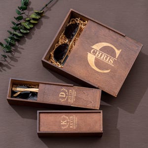 May include: Three wooden boxes with engraved initials and the word "Groomsman" on each. The boxes are brown and have a natural wood finish. The boxes are open and show the inside of the boxes, which are lined with brown paper. The boxes are all different sizes. The largest box has a pair of sunglasses inside.