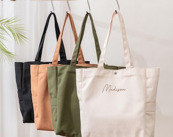 May include: Four canvas tote bags hanging from metal hooks on a wooden rod. The bags are black, tan, olive green, and cream. Each bag features two shoulder straps, side pockets, and a snap closure. The cream bag has the name Madison printed on it.