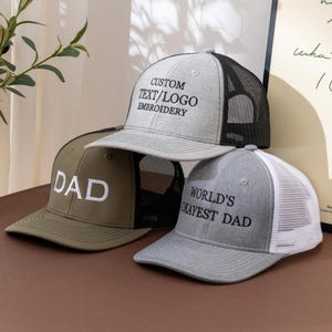 Può includere: Tre cappellini trucker con retro in rete. Il cappellino in alto è grigio mélange con rete nera e il testo CUSTOM TEXT/LOGO EMBROIDERY. Il cappellino a sinistra è verde oliva con rete nera e la parola DAD. Il cappellino a destra è grigio con rete bianca e WORLD'S OKAYEST DAD.