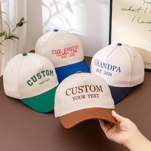 May include: Four baseball caps with off-white crowns and coloured brims. One has a green brim and says CUSTOM YOUR TEXT. One has a blue brim and says THE SMITHS Willow Creek EST. 2026. One has a dark blue brim and says GRANDPA EST. 2026. One has a brown brim.