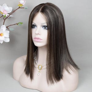 Op de afbeelding: Een bruine pruik met blonde highlights, gestyled met een middenscheiding en rechte, schouderlange lagen. De pruik wordt getoond op een mannequinhoofd, gecombineerd met een parelketting. De achtergrond is effen grijs.