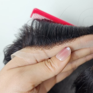 Puede incluir: Primer plano de una peluca negra con un frente de encaje. La peluca se sostiene en una mano con esmalte de u&ntilde;as rosa y un dise&ntilde;o de coraz&oacute;n blanco.