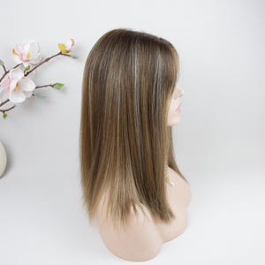 Op de afbeelding: Een pruik op een mannequinhoofd. De pruik heeft lang, steil haar met bruine en blonde highlights. Het haar is gestyled met een zijscheiding en valt tot op de schouders. Het mannequinhoofd heeft een neutrale huidskleur.