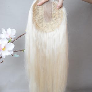 Pode incluir: Um topper de cabelo loiro com uma base de malha é segurado contra um fundo cinza. O cabelo é longo e liso, com uma textura natural. O topper foi projetado para adicionar volume e cobertura ao cabelo.