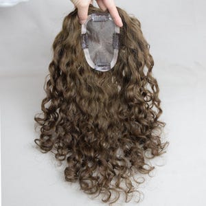 May include: A brown curly hair topper with a lace base and clips for attachment. The hairpiece is held up by a hand against a white background. The hair has a natural look and texture.