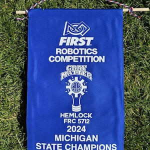 May include: Blue banner with white text, celebrating the FIRST Robotics Competition. The banner reads "FIRST Robotics Competition," "Gray Matter," "Hemlock FRC 5712 2024 Michigan State Champions." It is suspended by a wooden dowel and a red, white, and blue cord.