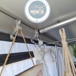 Könnte beinhalten: Innenansicht eines Wohnwagens mit Dekoration. Silberne Metallstangen und Halterungen halten weiße Vorhänge und eine Lichterkette mit Kugeln. Ein Holzschild hängt an einer Schnur. Ein Logo mit einer Wohnwagen-Illustration ist sichtbar.