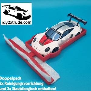 Może przedstawiać: Biało-czerwony zabawkowy samochód Porsche z czarnym spojlerem znajduje się w czerwonym urządzeniu czyszczącym, obok czerwono-białej podkładki czyszczącej. Obraz zawiera tekst "rdy2xtrude.com". Tło jest jednolicie niebieskie.