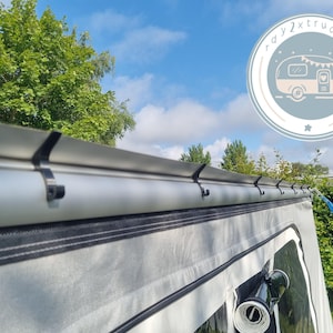 Könnte beinhalten: Ein Teil eines hellgrauen Campingzeltes mit silberfarbenem Dach und schwarzen Haken. Das Zelt ist im Freien unter blauem Himmel mit Bäumen im Hintergrund aufgebaut. Ein Logo mit einem Wohnwagen befindet sich in der oberen rechten Ecke.