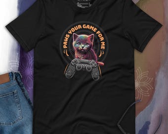 Puede incluir: Camiseta negra con una ilustración colorida de un gato sosteniendo un mando de videojuego. El texto "PAWS YOUR GAME FOR ME" está escrito en un círculo alrededor del gato.