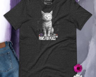 May include: Dark grey t-shirt with a smiling grey cat illustration and the text "PAWS FOR PEACE". The cat is surrounded by floral accents. The shirt is displayed with a pair of blue jeans and flowers.