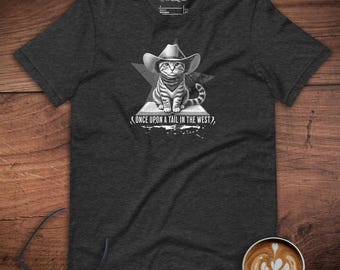 Pode incluir: Camiseta cinza escuro com uma ilustração em preto e branco de um gato usando um chapéu de cowboy e bandana. O gato está sentado em um triângulo, com o texto "ONCE UPON A TAIL IN THE WEST" abaixo.