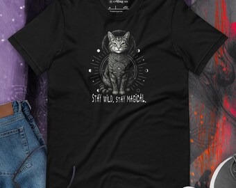 May include: Black t-shirt featuring a detailed illustration of a cat with a decorative collar, surrounded by celestial elements. The text "STAY WILD. STAY MAGICAL." is printed below the cat. The shirt is made of a soft material.