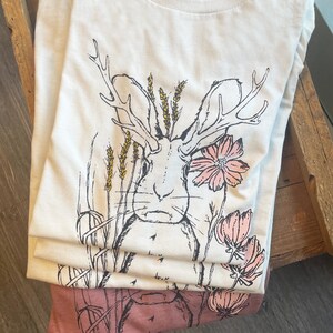 K&ouml;nnte beinhalten: Ein wei&szlig;es kurz&auml;rmeliges T-Shirt mit einer schwarzen Strichzeichnung eines Hasen mit Geweih, umgeben von gelben Weizen&auml;hren und rosa Blumen. Das T-Shirt hat einen Rundhalsausschnitt und ein sichtbares Bella Canvas Etikett am Innenkragen.