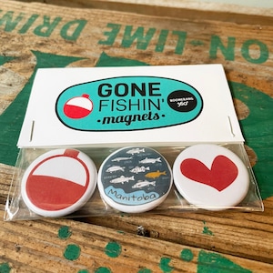Può includere: Un set di tre magneti rotondi in una confezione di plastica trasparente. I magneti presentano un galleggiante rosso e bianco, un disegno di pesce con la scritta "Manitoba" e un cuore rosso. La confezione recita "GONE FISHIN' magnets".