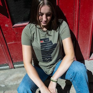 Könnte beinhalten: Eine Frau trägt ein grünes T-Shirt mit einer schwarzen Umrandung der Provinz Manitoba und dem Text "Ich liebe Prärien" in Schwarz. Sie trägt eine blaue Jeans und weiße Turnschuhe.