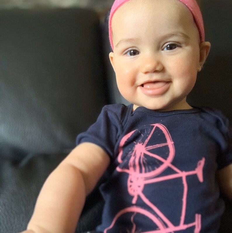 May include: A baby girl wearing a navy blue shirt with a pink bicycle graphic. She is wearing a pink headband and has a big smile on her face.