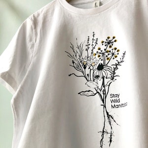Könnte beinhalten: Weißes T-Shirt mit einer schwarzen und goldenen Illustration von Wildblumen und dem Text "Stay Wild Manitoba".