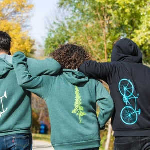 Op de afbeelding: Drie mensen dragen groene en zwarte hoodies met verschillende ontwerpen op de rug. De ontwerpen zijn een fiets, een boom en een fiets. De mensen lopen van de camera af.