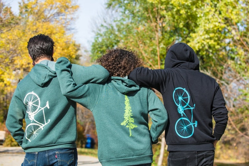 Op de afbeelding: Drie mensen dragen groene en zwarte hoodies met verschillende ontwerpen op de rug. De ontwerpen zijn een fiets, een boom en een fiets. De mensen lopen van de camera af.