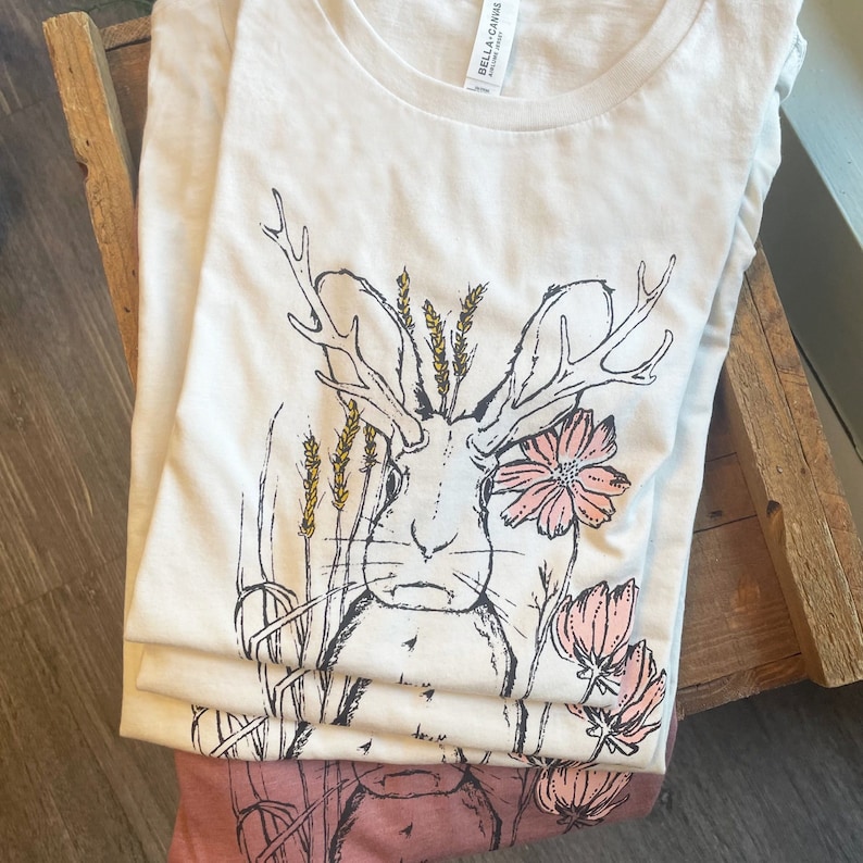 K&ouml;nnte beinhalten: Ein wei&szlig;es kurz&auml;rmeliges T-Shirt mit einer schwarzen Strichzeichnung eines Hasen mit Geweih, umgeben von gelben Weizen&auml;hren und rosa Blumen. Das T-Shirt hat einen Rundhalsausschnitt und ein sichtbares Bella Canvas Etikett am Innenkragen.