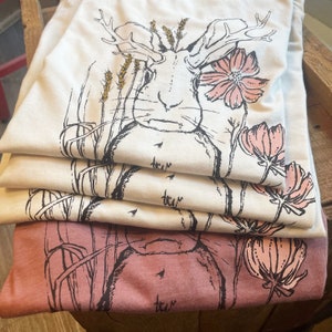 K&ouml;nnte beinhalten: Ein Stapel gefalteter T-Shirts mit einer schwarzen Strichzeichnung eines Hasen mit Geweih, Weizen&auml;hren und rosa Blumen. Die oberen drei T-Shirts sind cremefarben, w&auml;hrend das unterste T-Shirt in einem gedeckten Mauveton gehalten ist.