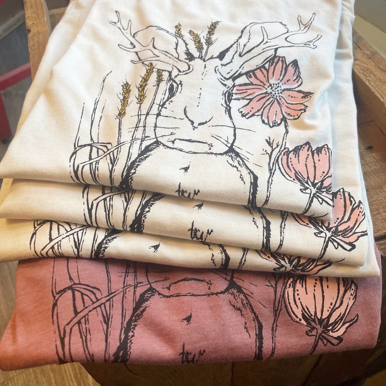 K&ouml;nnte beinhalten: Ein Stapel gefalteter T-Shirts mit einer schwarzen Strichzeichnung eines Hasen mit Geweih, Weizen&auml;hren und rosa Blumen. Die oberen drei T-Shirts sind cremefarben, w&auml;hrend das unterste T-Shirt in einem gedeckten Mauveton gehalten ist.