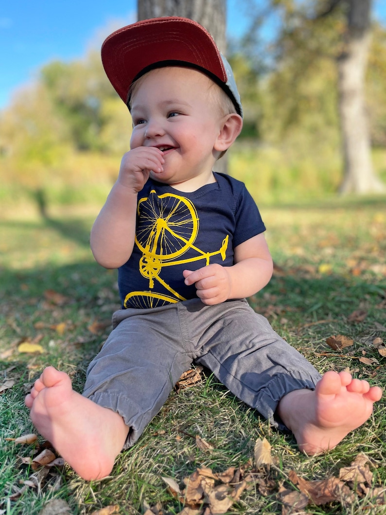 May include: A young child wearing a red and grey baseball cap sits on the grass, smiling and holding a finger to their mouth. They are wearing a navy blue shirt with a yellow bicycle graphic and grey pants.