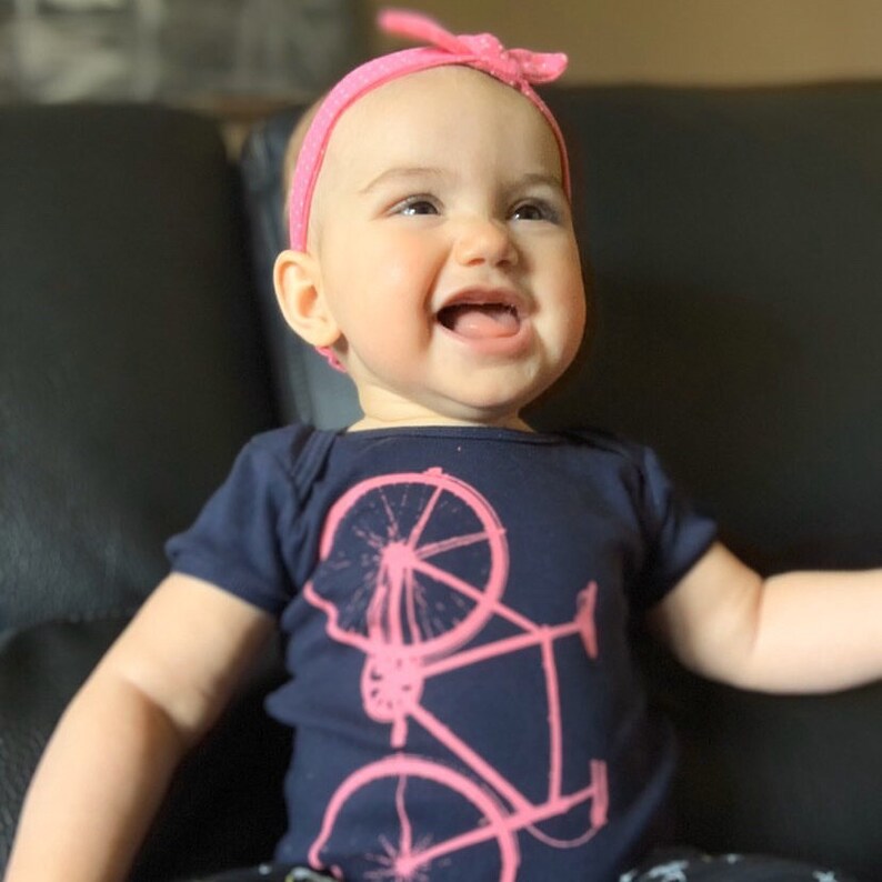 May include: A baby girl wearing a navy blue bodysuit with a pink bicycle graphic. She is smiling and wearing a pink headband.