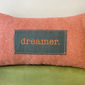 May include: A rust-coloured pillow with a dark green patch that says "dreamer." in orange lettering.