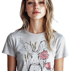 K&ouml;nnte beinhalten: Ein hellgraues Kurzarm-T-Shirt mit einer schwarzen Strichzeichnung eines Hasen mit Geweih, umgeben von gelben Weizen&auml;hren und drei rosa Bl&uuml;ten. Das Motiv ist mittig auf der Brust des Kleidungsst&uuml;cks platziert.