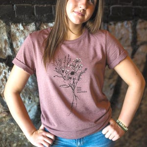 Könnte beinhalten: Ein malvenfarbenes Kurzarm-T-Shirt mit einer schwarzen Strichzeichnung eines Wildblumenstraußes mit Wurzeln. Der Text "Stay Wild Manitoba" ist in Schwarz neben dem Blumenmotiv aufgedruckt. Das Shirt hat einen Rundhalsausschnitt und umgekrempelte Ärmel.