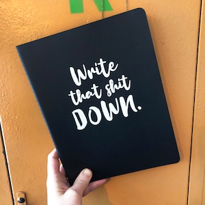 May include: Black notebook with the text "Write that shit DOWN." in white cursive lettering. The notebook is held by a person. The background is a yellow-orange locker.