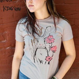 K&ouml;nnte beinhalten: Ein hellgraues Kurzarm-T-Shirt mit einer schwarzen Strichzeichnung eines Hasen mit langen Ohren. Der Hase ist von Weizen&auml;hren und drei rosa Bl&uuml;ten umgeben. Das Motiv befindet sich mittig auf der Vorderseite des Kleidungsst&uuml;cks.