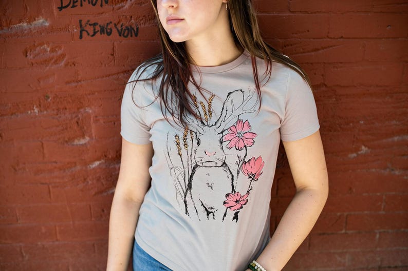 K&ouml;nnte beinhalten: Ein hellgraues Kurzarm-T-Shirt mit einer schwarzen Strichzeichnung eines Hasen mit langen Ohren. Der Hase ist von Weizen&auml;hren und drei rosa Bl&uuml;ten umgeben. Das Motiv befindet sich mittig auf der Vorderseite des Kleidungsst&uuml;cks.