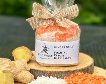 May include: A clear plastic bag tied with a brown ribbon contains a white and orange bath salt mixture. The bag has a white label that reads "SOAP STREET Handmade" and "GINGER SPICE FOAMING EPSOM BATH SALTS". There are pieces of ginger root on a wooden surface next to the bag.