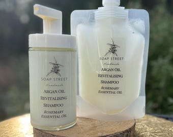 Può includere: Due bottiglie di shampoo rivitalizzante all'olio di argan con olio essenziale di rosmarino. Le bottiglie sono bianche con testo nero e una piccola immagine di una pianta. Le bottiglie sono appoggiate su una superficie di legno.